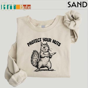 May include: Sand-colored sweatshirt with a black graphic of a squirrel holding a knife, with the text "PROTECT YOUR NUTS" above the squirrel. The sweatshirt has a crew neck and long sleeves. The brand name "Art Cute" is in the top left corner.