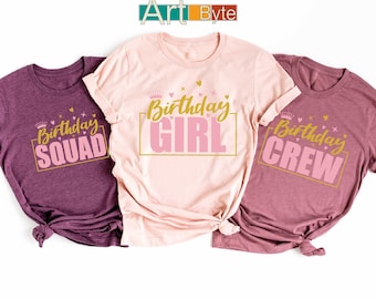 May include: Three short-sleeved t-shirts in shades of pink and purple. Each shirt has a different gold and pink graphic: "Birthday Squad", "Birthday Girl", and "Birthday Crew". The shirts are knotted at the bottom.