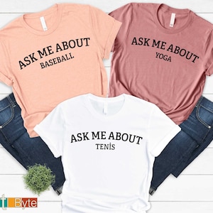 May include: Three t-shirts with the text "ASK ME ABOUT" above the sports. The shirts are peach, mauve, and white. The sports are baseball, yoga, and tennis. The shirts are folded with blue jeans.