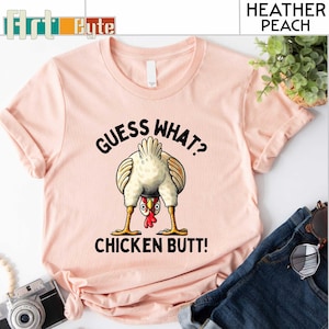 May include: Heather peach-colored t-shirt with the text "GUESS WHAT? CHICKEN BUTT!" and a cartoon chicken illustration. The shirt has short sleeves and a crew neck. The design features a chicken with its rear end facing the viewer.