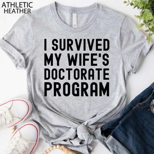 May include: Athletic heather gray t-shirt with the text "I SURVIVED MY WIFE'S DOCTORATE PROGRAM" in bold black letters. The shirt is knotted at the waist and paired with blue jeans and white sneakers.