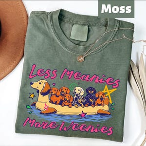 A green t-shirt with a graphic design featuring a group of dogs on a raft, along with the text "Less Meanies More Weenies" printed on it.