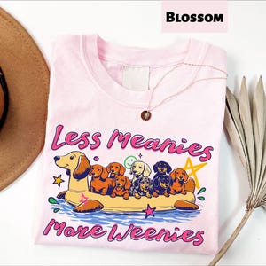 A pink t-shirt with a graphic design of a dog floating on a raft, surrounded by various dog breeds, and the text "Less Meany More Weenies" printed on it.