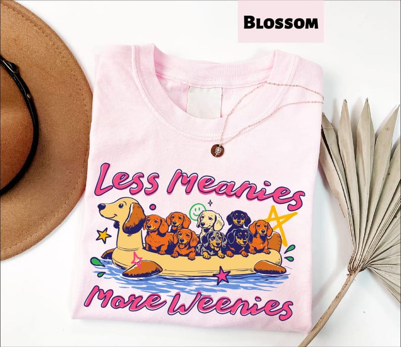 A pink t-shirt with a graphic design of a dog floating on a raft, surrounded by various dog breeds, and the text "Less Meany More Weenies" printed on it.