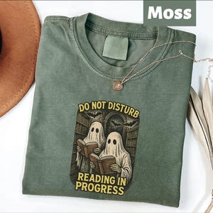 May include: A moss green crew neck t-shirt featuring a graphic of two ghosts wearing white sheets and reading books in a library. The text above the ghosts reads DO NOT DISTURB and the text below reads READING IN PROGRESS in yellow capital letters.