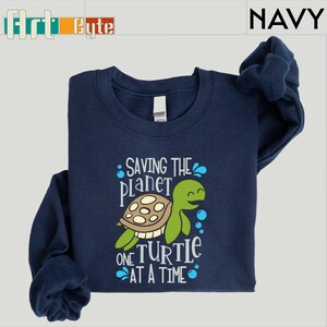 May include: Navy blue sweatshirt with a graphic of a smiling green turtle and the text "Saving the Planet One Turtle at a Time." The sweatshirt has long sleeves and a crew neck. The brand name "Flirt Cute" is in the top left corner.