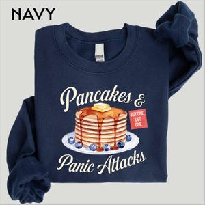 May include: Navy blue sweatshirt with the word "NAVY" at the top. The sweatshirt features a graphic of pancakes with butter and blueberries, and the text "Pancakes & Panic Attacks". A red square says "BUY ONE, GET ONE".