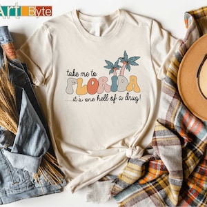 May include: Cream-colored t-shirt with the text "take me to FLORIDA...it's one hell of a drug!" in a retro font. The design includes palm trees. The shirt is styled with a denim jacket and a plaid scarf.