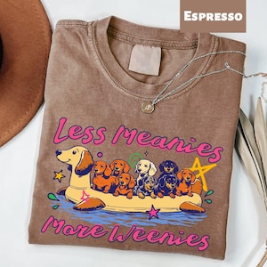 A t-shirt with a graphic design featuring a group of dogs in a boat, along with the text "Less Meany More Weenies" printed on it.