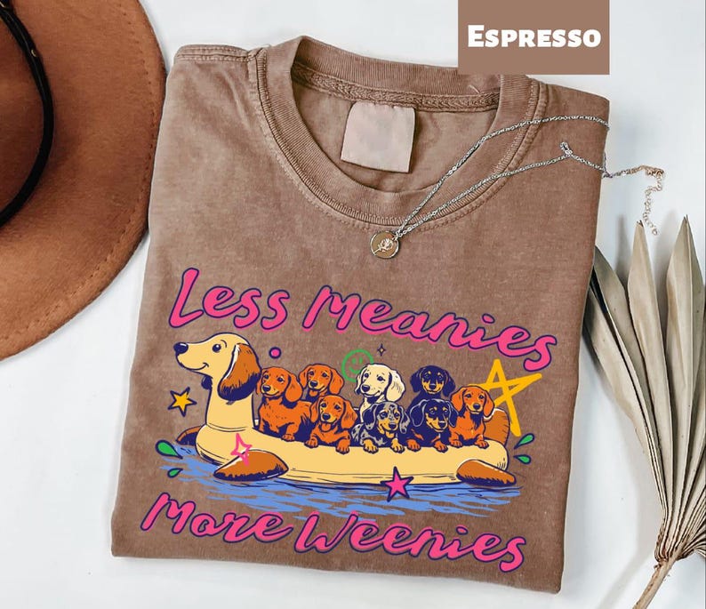 A t-shirt with a graphic design featuring a group of dogs in a boat, along with the text "Less Meany More Weenies" printed on it.