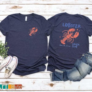 May include: Two navy blue t-shirts with a lobster design. One shirt features a small lobster on the front, the other has a larger lobster graphic with the words "Lobster Lover Club". White trainers and blue jeans are also in the image.