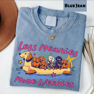 A blue t-shirt with a graphic design of a dog floating on a yellow float, surrounded by various cartoon-style dogs. The text on the t-shirt reads "Less Meany More Weenies".