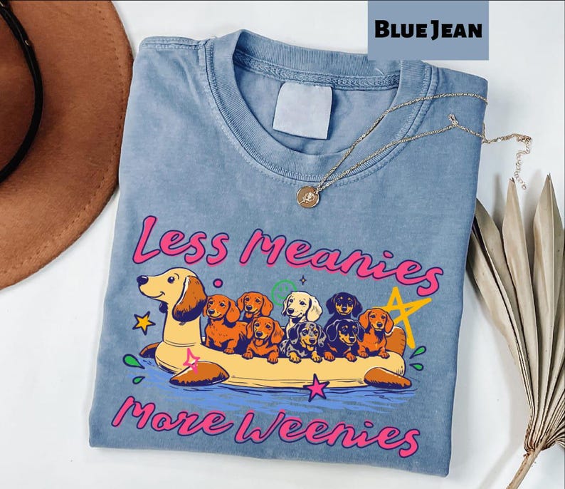A blue t-shirt with a graphic design of a dog floating on a yellow float, surrounded by various cartoon-style dogs. The text on the t-shirt reads "Less Meany More Weenies".