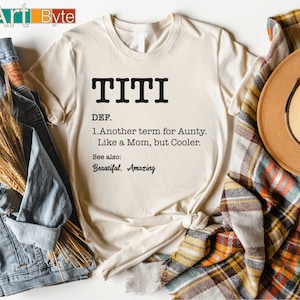 May include: A cream-coloured t-shirt featuring the word "TITI" in black, with a definition below. The definition reads "Another term for Aunty. Like a Mom, but Cooler." The shirt is styled with a plaid scarf and a brown hat.