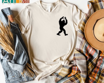 Puede incluir: Camiseta color crema con una silueta negra de Bigfoot formando un corazón con sus manos. La camiseta está anudada a la cintura. La marca "Art Byte" es visible en la esquina superior izquierda.