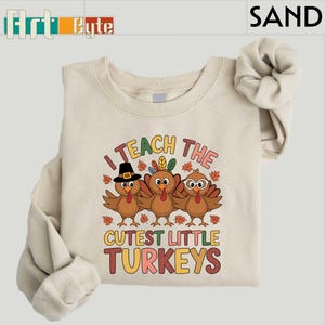 May include: A sand-colored sweatshirt with the text "I TEACH THE CUTEST LITTLE TURKEYS" in colorful letters. The graphic features three cartoon turkeys with festive accessories and autumn leaves. The sweatshirt has long sleeves.