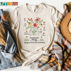May include: Cream-colored t-shirt with a floral design in a mason jar, featuring the words "Occupational Therapy" and "Helping You Grow To Your Full Potential." The shirt also lists areas of focus like ADLS, Play, and Fine Motor.
