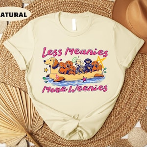 A white t-shirt with a graphic design featuring a group of dogs on a boat, accompanied by the text "Less Meany More Weenies" printed on it.