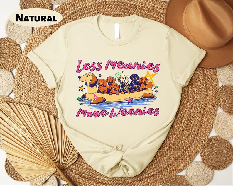 A white t-shirt with a graphic design featuring a group of dogs on a boat, accompanied by the text "Less Meany More Weenies" printed on it.