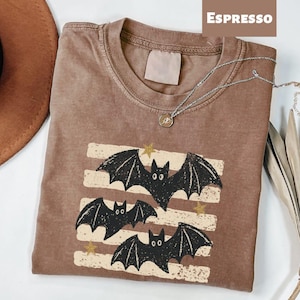 May include: An espresso-colored t-shirt featuring a graphic print of three black bats flying over horizontal distressed cream-colored stripes, accented with small gold stars. The shirt has a crew neckline and a tagless collar design.