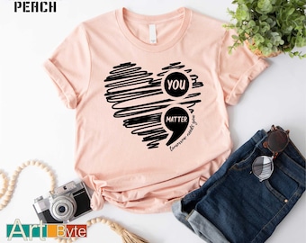Könnte beinhalten: Pfirsichfarbenes T-Shirt mit schwarzem Herzdesign. Das Herz besteht aus gekritzelten Linien und enthält die Worte "you matter" in einer Sprechblase. Der Text "tomorrow needs you" ist ebenfalls vorhanden. Das Shirt wird mit blauen Jeans kombiniert.