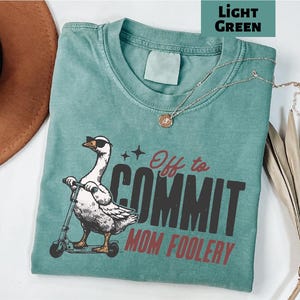 May include: A light green t-shirt with the phrase "Off to COMMIT MOM FOOLERY" and a graphic of a goose wearing sunglasses riding a scooter. A gold necklace is draped across the shirt. The shirt is folded on a white surface.