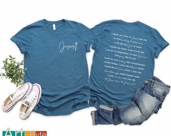 May include: Teal t-shirt with the word "Jesust" and a cross printed in white on the front. The back of the shirt features a prayer with white text. A pair of white sneakers and blue jeans are also in the image.