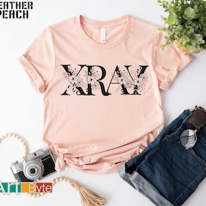 May include: A light peach-colored t-shirt with the word "XRAY" in black, adorned with floral designs. The shirt is styled with rolled-up sleeves and paired with blue jeans and sunglasses. The words "Heather Peach" and "Art Byte" are also visible.