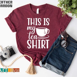 May include: Heather maroon t-shirt with the text "THIS IS my tea SHIRT" in white. The design includes a teacup and saucer graphic. The shirt is paired with denim shorts and accessories.