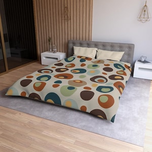 May include: A beige duvet cover with a repeating pattern of colorful, abstract shapes in shades of brown, orange, green, and blue. The bedding is on a gray bed with white pillows, and the room has wooden paneling.