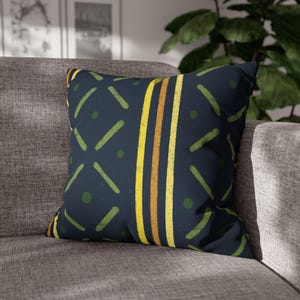May include: Square decorative pillow with a navy blue background and a geometric pattern. The pattern includes yellow, orange, and green vertical stripes, diagonal lines, and dots. The pillow is placed on a gray sofa.