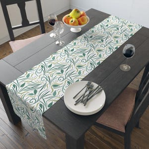 May include: A dining table set with a white and green patterned table runner. The table is set with plates, forks, wine glasses, and a bowl of apples. The table and chairs are dark wood.