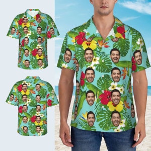 Puede incluir: Una camisa de manga corta de color azul claro con botones y un estampado tropical que presenta un patr&oacute;n repetido de un rostro de hombre, hojas verdes y flores rojas, amarillas y blancas.