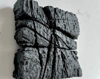 Könnte beinhalten: Eine dunkelgraue, strukturierte, rechteckige Skulptur mit rauer, steinartiger Oberfläche. Das Kunstwerk ist in Abschnitte mit sichtbaren Rissen und Linien unterteilt, wodurch eine fragmentierte Ästhetik entsteht. Das Stück ist an einer weißen Wand befestigt.