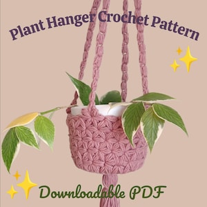 May include: A dusky pink crochet plant hanger with a white pot holding a green plant with white edges. The plant hanger has a starburst pattern and a tassel. Text reads "Plant Hanger Crochet Pattern" and "Downloadable PDF".
