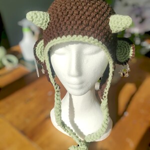 May include: A brown and green crocheted hat with a long green crocheted tie. The hat has two small green horns and two small green ears.
