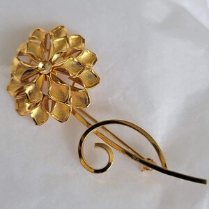 May include: A gold-toned flower brooch with a swirl design. The flower is made of many small petals and has a single round centre.
