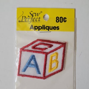 May include: An embroidered applique of a block with the letters A, B, and C on it. The applique is red, white and yellow. It is packaged on a white card with the words "Sew Perfect Appliques" and "80c" printed on it.