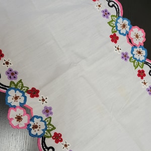 May include: White table runner with a pink and black scalloped edge. The edge is embroidered with colourful flowers in shades of red, pink, blue, green, and white.