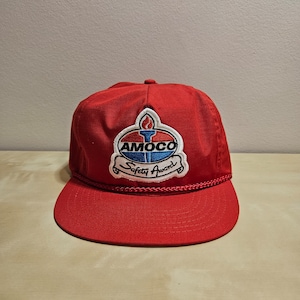 May include: Red baseball cap with a white and blue Amoco Safety Award patch. The patch features a flame and the Amoco logo.
