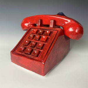 May include: A red ceramic telephone sculpture with a handset and a keypad. The phone is a solid, glossy red with a dark blue accent on the handset. The keypad has square buttons. The sculpture is on a white surface.