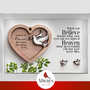 May include: A wooden heart-shaped ornament with a dove cutout and the text "Feathers are Reminders that angels are always near". The ornament is in a white box with a red cardinal logo and the text "Always Near". Two small dove earrings are also included.