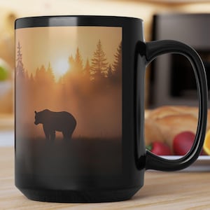 Könnte beinhalten: Schwarze Keramik-Tasse mit einem Bären-Silhouette-Design in einem Wald bei Sonnenuntergang. Das Design zeigt einen Bären, der in einem Feld mit Bäumen und einer hellen Sonne steht. Die Tasse hat einen großen Griff.