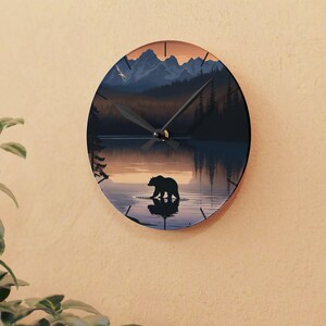 May include: Circular wall clock depicting a tranquil scene of a bear in a lake, with mountains and trees. The clock face has a dark blue and orange colour scheme, with black clock hands. The clock is mounted on a beige wall.