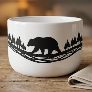 May include: A white ceramic mug featuring a black silhouette graphic of a bear walking through a forest of pine trees. The design wraps around the centre of the mug, depicting the bear in profile against a backdrop of trees and abstract lines.