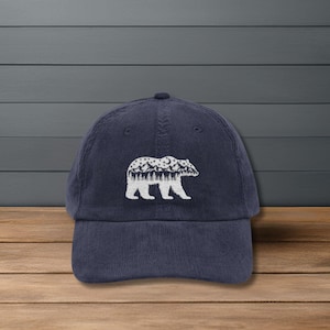 Könnte beinhalten: Marineblaue Cord-Baseballkappe mit weißer gestickter Bärensilhouette. Das Bären-Design umfasst eine Bergkette, Mond und Sterne. Die Kappe hat einen gebogenen Schirm und einen verstellbaren Riemen.