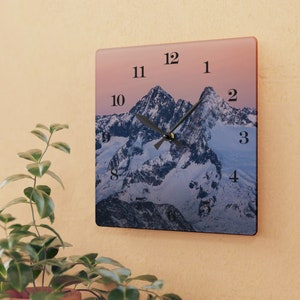 May include: A square clock featuring a mountain scene with a pink sky. The clock face has black numbers and hands. The numbers 1, 2, 3, 4, 8, 9, 10, 11, and 12 are visible.