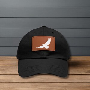 May include: Black baseball cap with a brown leather patch featuring a white eagle design. The cap has a curved brim and a classic, adjustable fit. The eagle graphic is a silhouette of a bird in flight.
