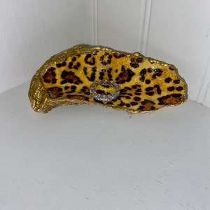 May include: A gold and brown leopard print oyster shell ring dish. A silver ring is sitting in the dish.