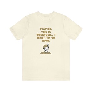 May include: A beige t-shirt with a gold graphic of a soldier with the text "STATION. THIS IS OBSERVER... I WANT TO GO HOME" printed on the front.